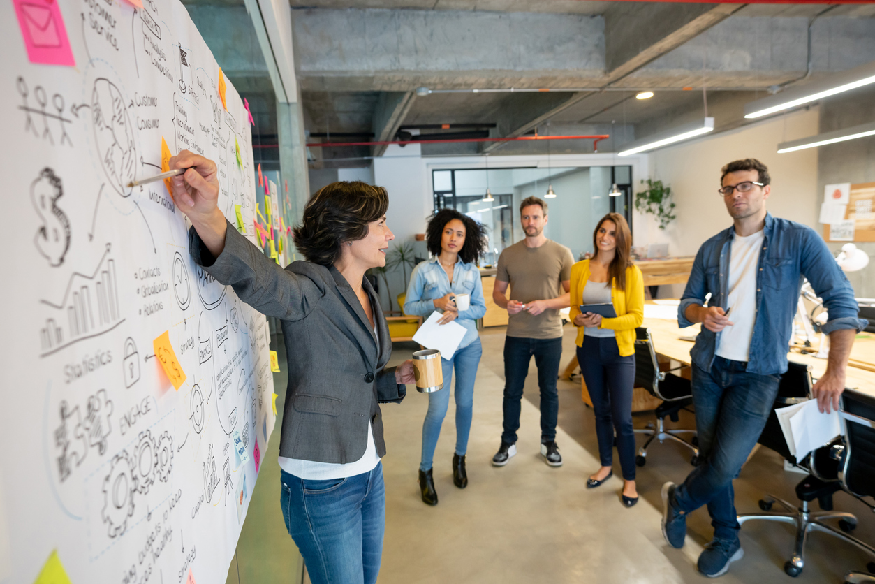 Latin American woman makes a business presentation during a meeting in a creative office, pointing out her business plan to her team.